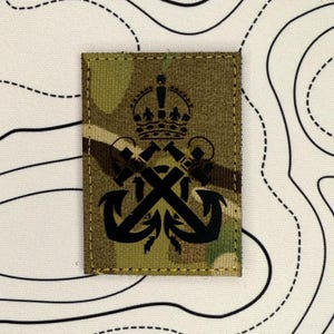 May include: A rectangular patch with a camouflage pattern. The patch features a black emblem with a crown, crossed cannons, and anchors. The patch is likely a military or nautical insignia.
