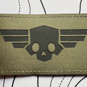 Puede incluir: Parche rectangular verde oliva con un diseño de calavera y alas negras. El parche tiene un borde cosido y está hecho de una tela texturizada. La calavera tiene dos cuencas de los ojos y una nariz. Las alas están estilizadas.