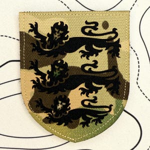 Könnte beinhalten: Ein schildförmiger Aufnäher mit Tarnmuster. Auf dem Aufnäher sind drei schwarze Löwen abgebildet. Der Aufnäher ist mit einem hellbraunen Stoff eingefasst und hat einen genähten Rand.