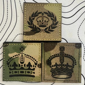 May include: Three square patches with a camouflage pattern. Each patch features a black crown design. One patch has a crown within a laurel wreath. The patches are stitched with a tan thread.