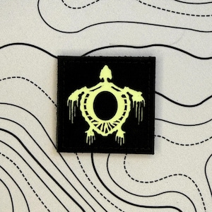 May include: A black square patch with a neon green turtle design. The turtle has a circular body, detailed shell and dripping accents. The patch is set against white fabric with black contour lines.