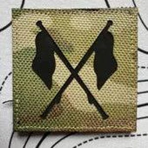 Könnte beinhalten: Quadratischer Aufnäher mit Tarnhintergrund und einem schwarzen gekreuzten Flaggen-Design. Der Aufnäher hat ein strukturiertes Aussehen und einen genähten Rand. Die Flaggen sind stilisiert und scheinen zu wehen. Das Gesamtdesign ist einfach und kühn.