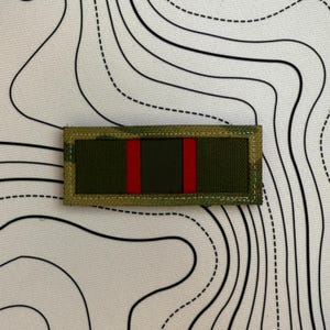 Puede incluir: Un parche rectangular con borde de camuflaje, un rectángulo central verde oscuro y dos franjas rojas verticales. El parche es probablemente para uso militar o táctico.