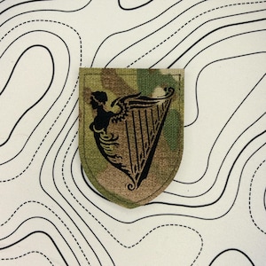 以下が含まれることがあります： 黒いハープと女性のシルエットが描かれた迷彩パッチ。シールド型のパッチには、ハープと女性の横顔の詳細なデザインが施されています。背景には地形図パターンがあります。