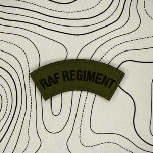 Può includere: Una toppa verde oliva con la scritta nera "RAF REGIMENT", arcuata verso l'alto. La toppa è posta su uno sfondo bianco con un disegno di mappa topografica nera.