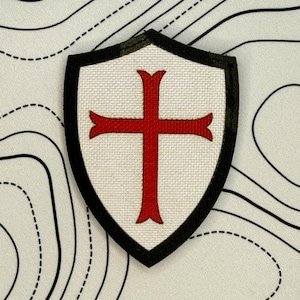 Puede incluir: Parche en forma de escudo con fondo blanco y diseño de cruz roja. El parche está delineado en negro y tiene una apariencia texturizada. Es probable que el parche sea un emblema o insignia.