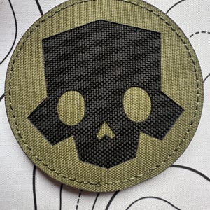 Pode incluir: Um patch circular com um desenho de caveira preta sobre um fundo verde-oliva. A caveira tem duas órbitas oculares circulares, um nariz triangular e uma cabeça retangular. O patch é bordado com costuras verde-oliva.