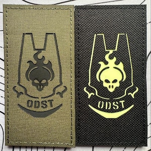 May include: Two rectangular patches. One is olive green with a black emblem featuring a skull and the letters ODST. The other is black with a glowing yellow emblem of the same design.