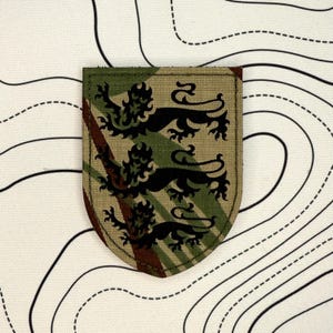 May include: A shield-shaped patch with a camouflage pattern in green, brown, and tan. The patch features three black heraldic lions. The background is a white fabric with black contour lines.