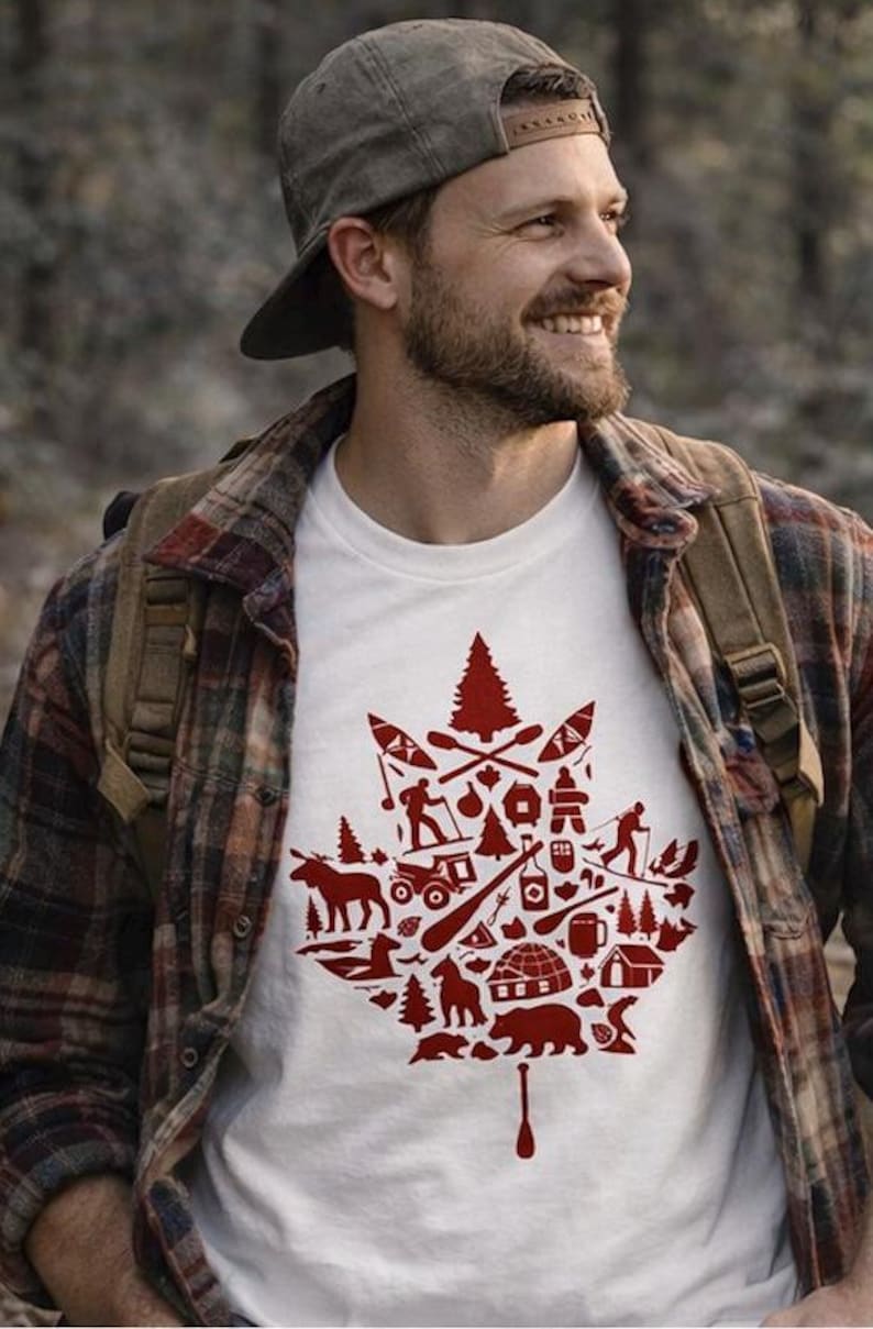 Peut inclure: T-shirt blanc avec un motif de feuille d'érable rouge rempli d'illustrations sur le thème du plein air. Le t-shirt est porté par une personne portant une chemise à carreaux et une casquette. Le motif comprend des images de canoës, d'arbres et d'animaux sauvages.