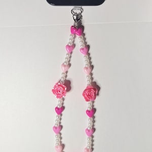 May include: A pink and white beaded phone charm. The charm features a string of white pearls, pink hearts, and pink rose-shaped beads. The charm is attached to a silver-colored clasp.