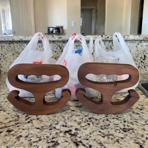 Puede incluir: Dos soportes de bolsas de madera en acabado marrón oscuro. Cada soporte tiene un diseño único en forma de ancla con un asa y una base curva. Los soportes se muestran sobre una encimera moteada, con bolsas de plástico en el fondo.