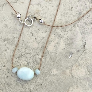 May include: A delicate necklace featuring a light blue, oval-shaped stone pendant, flanked by two smaller stones. The necklace is strung on a thin, tan cord with a silver clasp. The stones have a smooth, polished appearance.