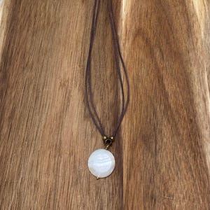 May include: A delicate necklace featuring a round, white pendant with a subtle iridescent sheen. The pendant is suspended from a brown cord, connected with a small, decorative clasp. The necklace rests on a wooden surface.