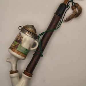 May include: A vintage tobacco pipe with a white ceramic bowl and a wooden stem. The bowl is decorated with a painted scene and a metal lid. The pipe has a curved design and is accented with green string and tassels.