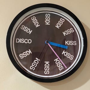 May include: A round wall clock with a black frame and a dark gray face. The clock face features the word "DISCO" and the word "KISS" repeated in white. The hour and minute hands are blue and pink.