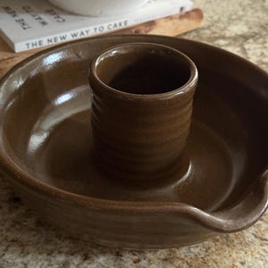 May include: A brown ceramic chicken roaster with a central cup for the chicken and a surrounding dish. The roaster has a glossy finish and is designed for cooking poultry. The design is simple and functional.
