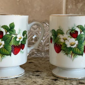 May include: Two white ceramic mugs with a gold rim and a pedestal base. Each mug is decorated with a strawberry and floral design. The mugs have a curved handle.