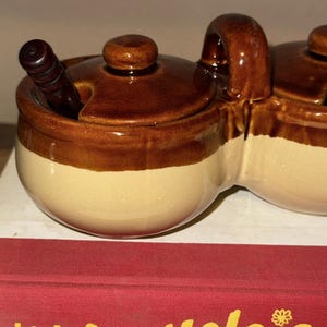 May include: A two-section ceramic condiment server with a brown glaze on the top and a cream-colored base. Each section has a lid and a handle. One section has a small wooden spoon.