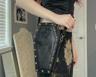 Könnte beinhalten: Eine schwarze Umhängetasche in Sargform mit einem geprägten Rosenmotiv auf der Vorderseite. Die Tasche ist an den Rändern mit silberfarbenen Metallnieten verziert und verfügt über einen schwarzen Schulterriemen mit silbernen Beschlägen.