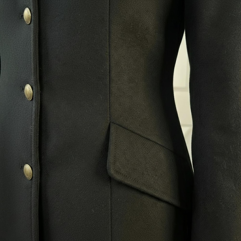 K&ouml;nnte beinhalten: Nahaufnahme eines schwarzen Blazers mit drei goldenen Kn&ouml;pfen auf der linken Seite. Der Blazer hat eine taillierte Passform und eine Tasche auf der rechten Seite. Der Stoff scheint ein glattes, gewebtes Material zu sein.