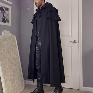 May include: A floor-length black hooded cloak featuring a layered shoulder capelet design. The garment includes a front tie closure with long fabric ribbons and vertical decorative stitching along the front edges, worn over black trousers and shoes.