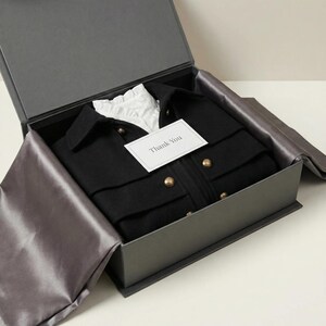 K&ouml;nnte beinhalten: Eine schwarze Jacke mit goldenen Kn&ouml;pfen und ein wei&szlig;es Hemd mit Kragen werden in einer grauen Geschenkbox pr&auml;sentiert. Die Jacke ist ordentlich gefaltet, und eine kleine Karte mit "Thank You" liegt obenauf. Die Box ist mit grauem Satin ausgekleidet.
