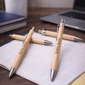 Peut inclure: Trois stylos à bille rétractables avec des corps en bois clair et des pointes, clips et boutons-poussoirs en métal argenté. Chaque stylo présente le texte imprimé en noir "Votre texte" sur le côté du corps en bois.