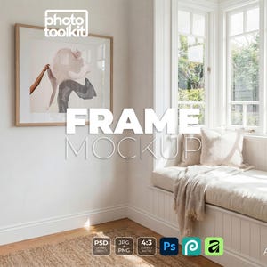 May include: A square wooden picture frame hanging on a white wall, containing an abstract print with a tan and dark gray color palette. The frame is positioned above a window seat with a beige cushion and a throw blanket in a bright room.