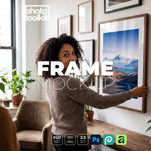 May include: A woman is hanging a framed landscape print on a white wall. The frame is brown with a white mount. The image shows a mountain and road scene. The text "FRAME MOCKUP" is overlaid. Other framed art pieces are visible.
