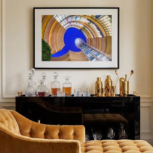 May include: A framed architectural photograph hangs above a black bar cart. The cart displays decanters, glassware, and gold bar tools. A tufted, mustard-colored chaise lounge sits in the foreground, complementing the room's decor.