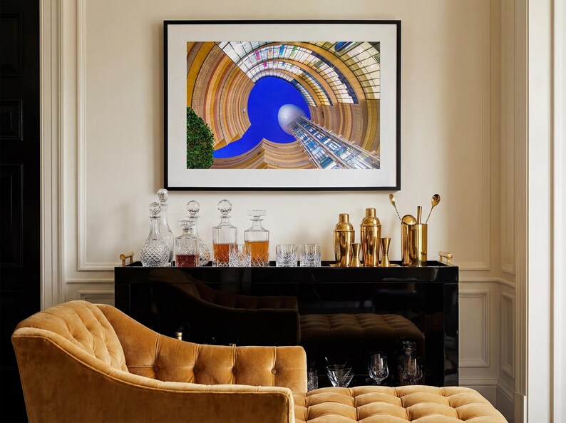 May include: A framed architectural photograph hangs above a black bar cart. The cart displays decanters, glassware, and gold bar tools. A tufted, mustard-colored chaise lounge sits in the foreground, complementing the room's decor.