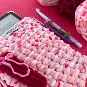 May include: A handmade, pink and white crocheted item with red accents, possibly a tablet case, lies on a pink surface. A small, dark red crocheted ballet slipper charm is attached. Two crochet hooks and balls of yarn in pink and burgundy are also visible.