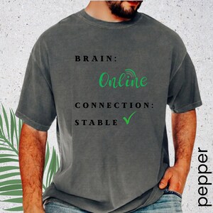 May include: A charcoal gray short-sleeve t-shirt featuring black text that reads BRAIN: followed by the word Online in green script, then CONNECTION: and STABLE with a green checkmark symbol. The shirt has a crew neckline and a relaxed fit.