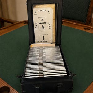 May include: A black metal bridge card case with a handle, containing a stack of silver-edged cards. The open lid displays a white card with black text reading SECTION A, NORTH 1, SOUTH 1, EAST, and WEST, along with other printed instructions.