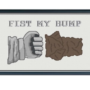 May include: Cross-stitch art with the text "FIST MY BUMP" above a gray fist and a brown shape. The artwork is framed in a dark blue frame, set against a light background.