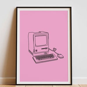 Puede incluir: Impresión enmarcada con un dibujo lineal negro de un ordenador retro sobre un fondo rosa. La imagen muestra un monitor, un teclado y un ratón, con el cable del ratón formando un corazón. La impresión se muestra sobre un suelo de madera.