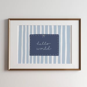 May include: A framed print with a blue and white striped background. A navy blue rectangle with the words "hello world" in cursive is centered on the print. A brown lamp with a beige lampshade sits on a wooden cabinet.