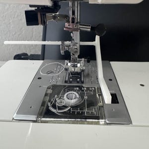 Pode incluir: Close-up da agulha e do calcador de uma máquina de costura. A máquina é branca com uma agulha prateada e uma placa de plástico transparente com medidas em centímetros. Uma guia de plástico branco está anexada.