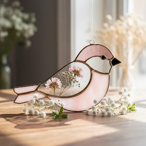 Könnte beinhalten: Ein dekorativer Vogel aus Glas in Rosa und Klarglas. Der Vogelkörper enthält getrocknete Blumen. Er hat einen schwarzen Schnabel und hängt an einer Schnur. Der Vogel sitzt auf einer Holzoberfläche. Er ist etwa 15 cm lang.