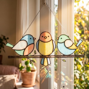 May include: A stained glass window decoration featuring three colorful birds perched on a branch. The birds are in shades of blue, pink, yellow, and green, with a metal chain for hanging. The window art is displayed in front of a window.