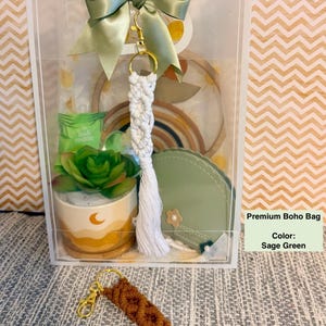 May include: A clear box with a sage green bow and a macrame keychain. Inside are a small succulent, a green pouch, and a rainbow decoration. The text "Premium Boho Bag" and "Color: Sage Green" are visible.