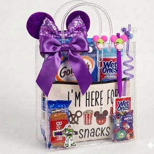 May include: A clear tote bag filled with snacks and Disney-themed accessories. The bag features a purple bow and mouse ears. Inside are snacks, Wet Ones wipes, and a pen. The tote bag has the text "I'M HERE FOR snacks".