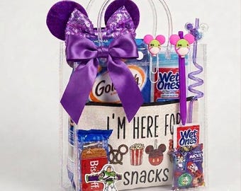 Puede incluir: Una bolsa de mano transparente llena de bocadillos y accesorios temáticos de Disney. La bolsa presenta un lazo morado y orejas de ratón. En el interior hay bocadillos, toallitas húmedas Wet Ones y un bolígrafo. La bolsa de mano tiene el texto "I'M HERE FOR snacks".