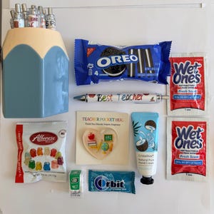 Peut inclure: Un porte-crayons bleu et cr&egrave;me contenant des stylos, un paquet de biscuits Oreo, deux sachets de lingettes antibact&eacute;riennes Wet Ones, un stylo Best Teacher, un jeton en forme de c&oelig;ur Teacher Pocket Hug, un tube de cr&egrave;me pour les mains Cristalbox &agrave; la noix de coco, un paquet d'oursons en g&eacute;latine Albanese, une petite bo&icirc;te de Tic Tac et un paquet de chewing-gum Orbit &agrave; la menthe.