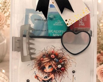 Puede incluir: Una caja de regalo transparente atada con un lazo negro, que contiene productos de belleza y una bolsa decorativa. La bolsa presenta una ilustración de una niña con cabello floral. Otros artículos incluyen una pinza para el pelo, un espejo en forma de corazón y botellas de productos para el cuidado corporal.