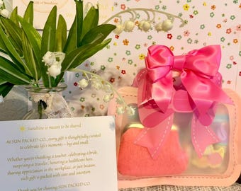 Puede incluir: Un juego de regalo rosa en una funda transparente, atado con un gran lazo rosa. El juego incluye un pequeño bolso rosa y otros artículos. El fondo presenta papel con estampado floral y un jarrón de lirios de los valles.