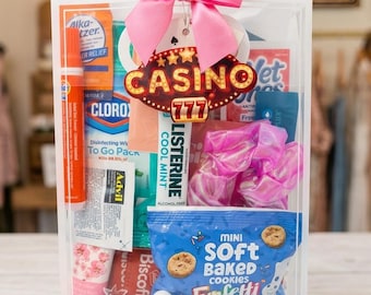 Puede incluir: Una bolsa de regalo transparente llena de varios artículos, incluyendo snacks, artículos de tocador y un lazo rosa. Una etiqueta con temática de casino con el número 777 cuelga del lazo. Fichas de póker y naipes son visibles en la parte inferior.