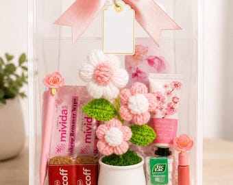 Puede incluir: Una caja de regalo transparente atada con un lazo de satén rosa, llena de productos de belleza y golosinas. Incluye toallitas desmaquillantes rosas, galletas Biscoff, una pequeña planta en maceta con flores de ganchillo y crema de manos de flor de cerezo.