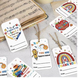 Peut inclure: Une collection d'&eacute;tiquettes cadeaux rectangulaires blanches orn&eacute;es d'illustrations color&eacute;es sur le th&egrave;me de l'&eacute;cole, incluant des arcs-en-ciel, des pommes, des crayons et des livres. Chaque &eacute;tiquette poss&egrave;de un trou avec de la ficelle, du texte imprim&eacute; comme "Thank You" ou "Best Teacher Ever" et des lignes pour "To" et "From".